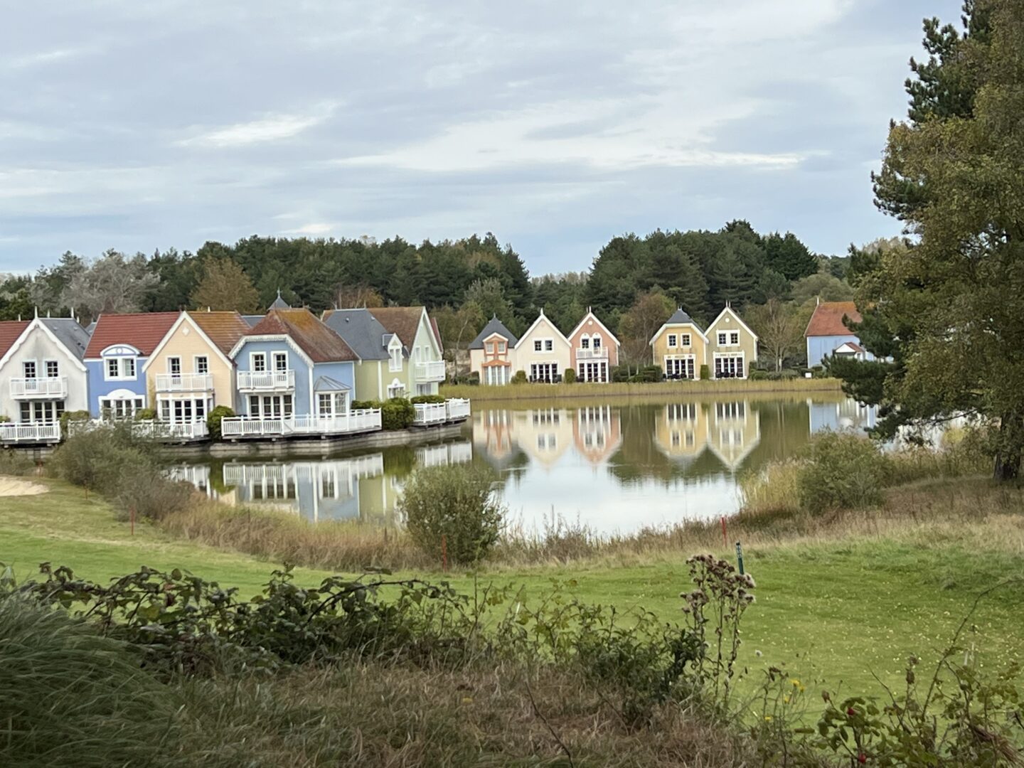 Maisons colorées alignées au bord d’un lac, avec leurs reflets dans l’eau et une forêt en arrière-plan au village de Belle Dune